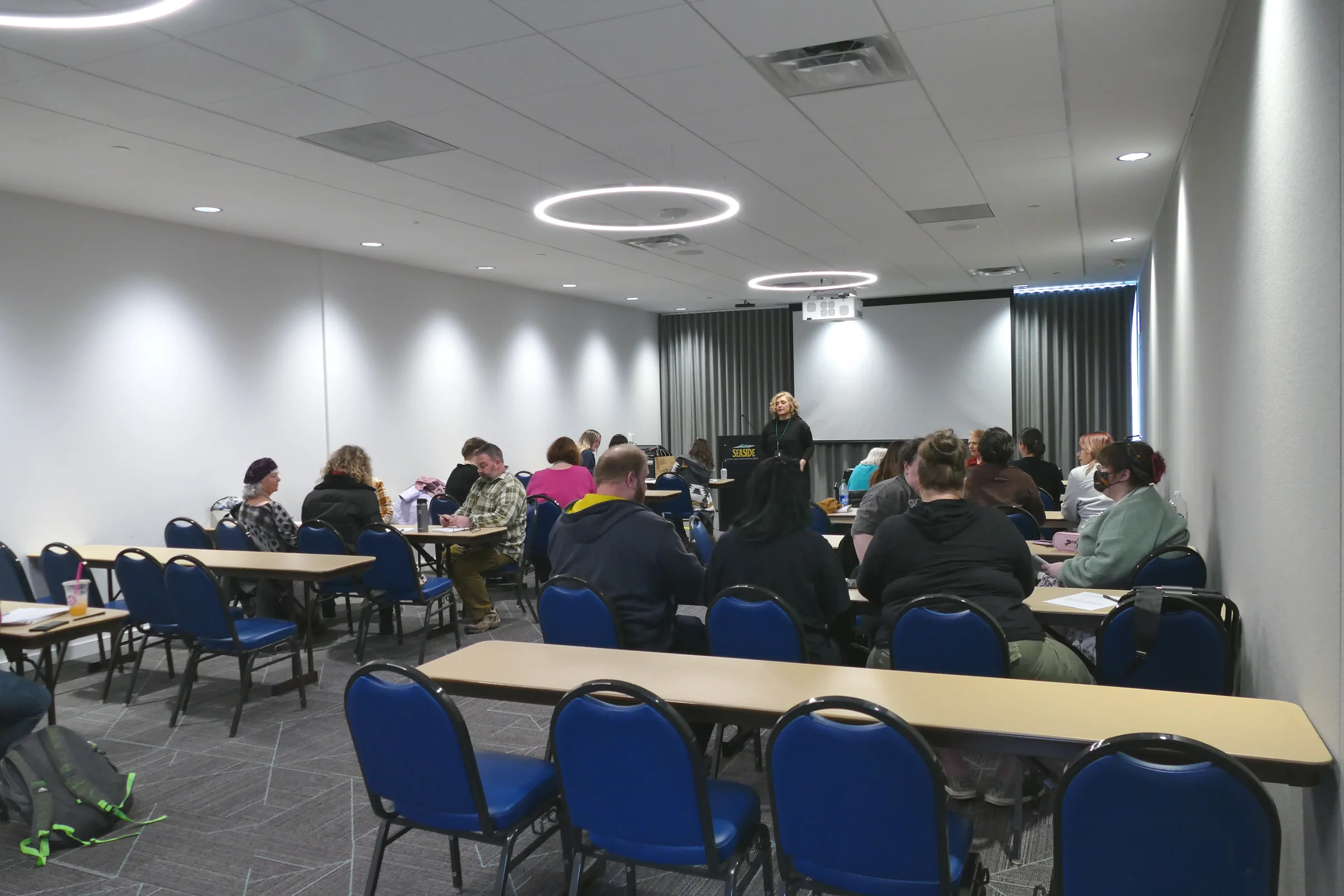 An instructor speaking at the podium to a full classroom of seated attendees taking notes